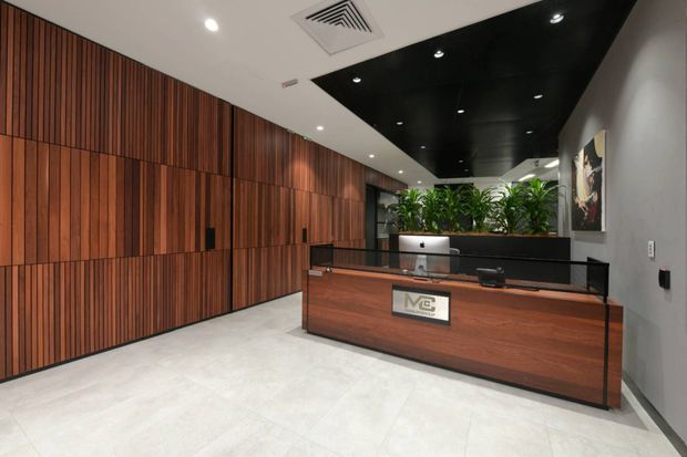 A contemporary commercial reception area with a large timber-panelled feature wall, a dark wood reception desk with glass screen, integrated computer workstation, indoor plants, and a black recessed ceiling feature with downlights. The space has a polished light stone or porcelain floor and a refined, professional atmosphere.