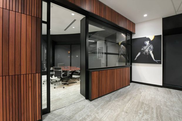 A contemporary commercial office interior with a glazed meeting room enclosed in black-framed glass, bordered by vertical timber wall panelling and matching lower joinery. The meeting room contains a conference table and office chairs. Light patterned carpet, white ceiling panels with recessed downlights, black accents, and monochrome wall artwork create a refined professional workspace.