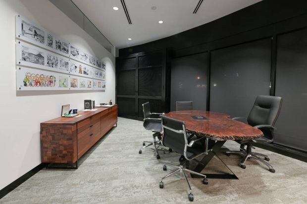 A modern executive meeting room with a large round reddish wood conference table surrounded by black leather office chairs. A long timber sideboard with drawers sits against a white wall displaying framed architectural sketches, while the opposite wall features dark full-height panelled joinery or glazing. Recessed ceiling lighting, linear air vents and grey patterned carpet create a refined corporate interior.