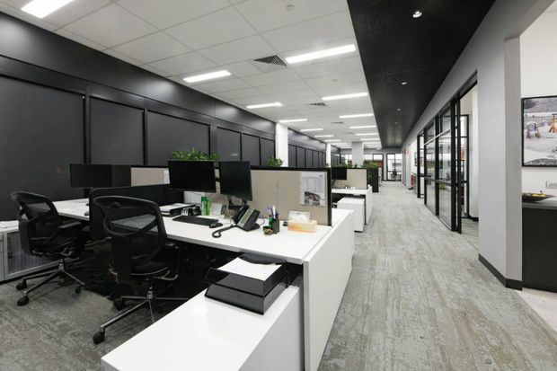 A modern open-plan commercial office interior with rows of white workstations, black mesh office chairs, desktop monitors, telephones and low planter dividers. The space has dark charcoal feature walls and ceiling panels, white structural columns, extensive glazed meeting-room partitions on the right, recessed linear office lighting, and grey patterned carpet flooring. The long corridor and repeated workstations create a spacious, organised workplace environment.