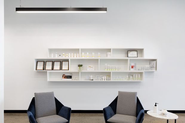 Minimalist consultation or reception area with two upholstered armchairs, a small round side table, and floating white display shelves holding framed certificates and product containers. The room has white walls, a dark floor trim, neutral carpet, and a suspended linear pendant light.
