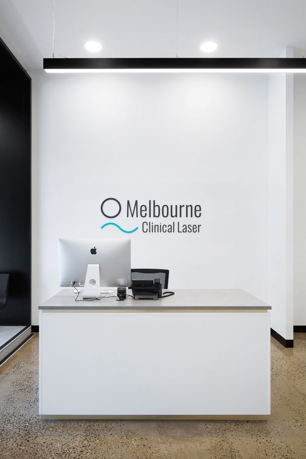 A minimalist clinical reception area featuring a white reception desk with a light grey benchtop, computer workstation and printer. The wall behind displays the Melbourne Clinical Laser logo, while a black linear pendant light, recessed ceiling lights, dark wall elements and polished exposed aggregate flooring create a clean, contemporary medical-commercial interior.
