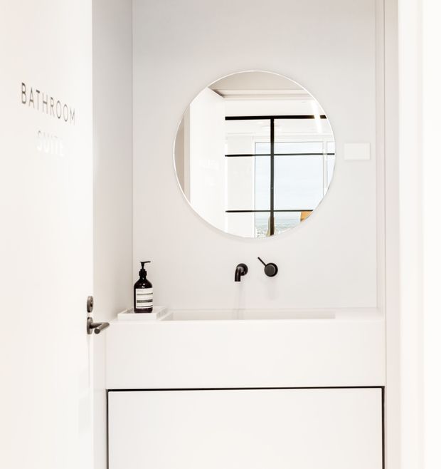 Compact minimalist bathroom vanity with a white integrated basin and flat-front cabinetry, matte black wall-mounted tapware and round mirror. A partially open door is labeled “BATHROOM SUITE,” and the mirror reflects a bright interior with black-framed glazing.