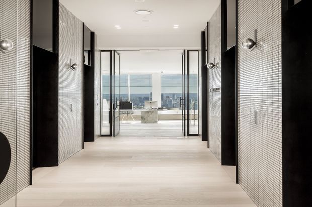 A contemporary interior corridor or entry passage with pale timber flooring, textured light mosaic wall surfaces, and dramatic black vertical door and wall elements. Glazed doors open toward a bright living or kitchen area with floor-to-ceiling windows and a distant urban view. Recessed ceiling lighting and minimal detailing create a refined, high-contrast atmosphere.
