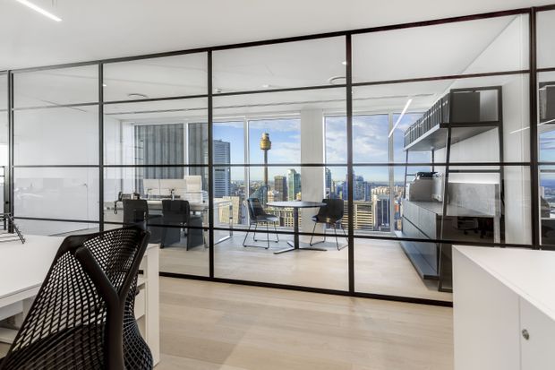A bright contemporary apartment interior with pale timber flooring, full-height black-framed glass partitions, and an open-plan workspace or living area overlooking a dense city skyline. A compact desk with chairs is visible behind the glazing, while dark built-in shelving and cabinetry occupy the right side. The space has clean lines, abundant daylight, and a refined urban feel.