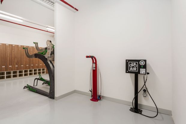 Clean, bright commercial or institutional fitness facility interior with white walls, polished light-grey flooring, exposed services, and red-painted pipework. A bicycle air pump station is mounted on the wall in the foreground, while exercise equipment and extensive wooden locker storage are visible in the adjoining area.