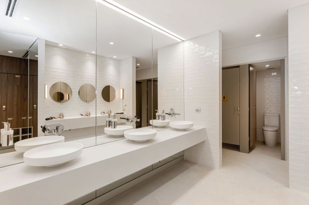 A bright, contemporary commercial-style bathroom or powder room with a long white vanity, multiple round vessel basins, wall-mounted taps, large mirror panels, and circular mirrors. White textured wall finishes, pale flooring, recessed lighting, and an open toilet cubicle area create a clean, spacious fit-out.