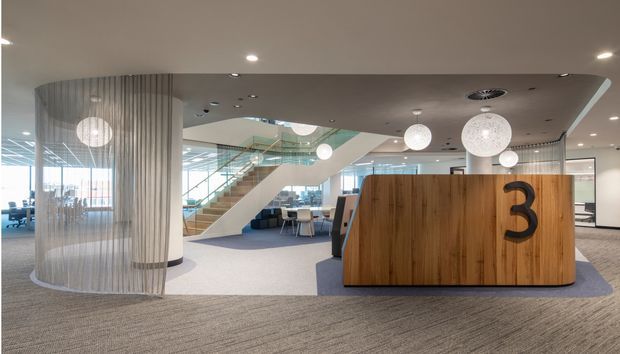 Contemporary commercial interior lobby or reception area with a large timber reception desk marked with the number 3, patterned carpet, spherical pendant lights, a translucent curved screen, glass balustrades, and a prominent staircase leading to an upper level. The space is open, bright, and professionally fitted out.