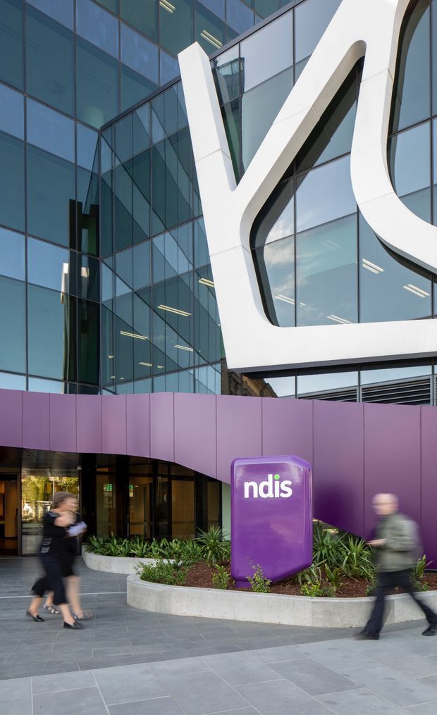 Exterior view of a contemporary multi-storey commercial building with a reflective glass curtain wall, bold white sculptural façade framing, and a curved purple entrance canopy. A purple NDIA/ndis sign stands in a landscaped planter at the pedestrian forecourt, with people walking past.