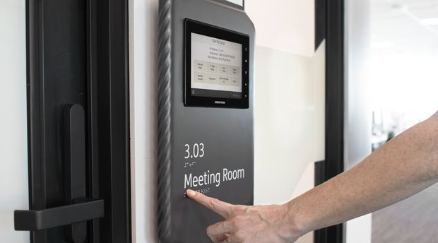 Close-up of a modern office meeting-room entrance with a dark-framed glass door, a wall-mounted digital room-booking touchscreen, and clear white wayfinding text reading “3.03 Meeting Room.” A person is touching the screen; a bright office corridor or workspace is visible beyond.