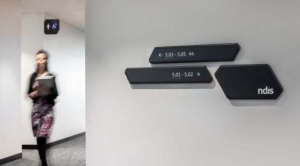 Interior wayfinding signage in a clean commercial corridor. Dark, angled wall-mounted signs direct visitors to rooms 5.01–5.05 and identify an NDIS area, with accessible toilet signage above a blurred person in the hallway. The space has white walls, a light ceiling, and grey carpet.