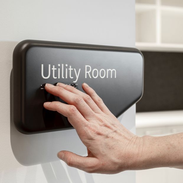 Close-up of a hand operating a black wall-mounted touch panel labelled “Utility Room,” with tactile dots beneath the text. The panel is mounted on a light wall, with a softly blurred glimpse of white cabinetry and a dark benchtop or backsplash in the background.