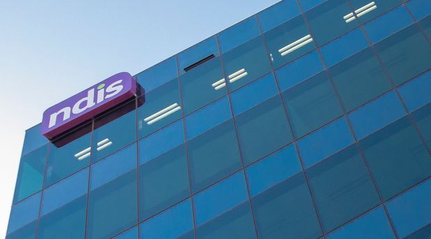 Low-angle exterior view of a modern commercial building with a blue reflective glass curtain-wall facade and a prominent purple building sign reading “ndis.”