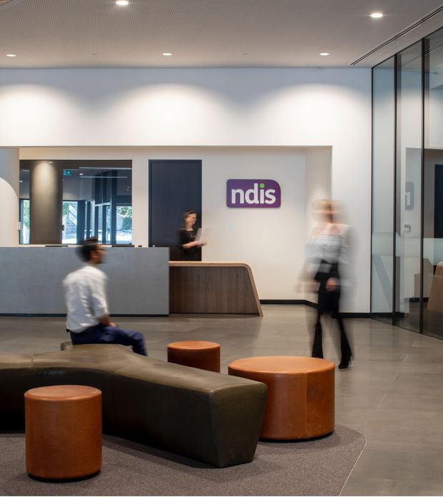 A contemporary commercial reception and waiting area with a curved timber-fronted reception desk, dark upholstered seating, cylindrical leather ottomans, glass-partitioned offices, polished flooring, and a visible NDIS sign. The space has a ribbed acoustic ceiling, recessed downlights, and a neutral grey, white, timber, and brown palette. Motion-blurred people are present in the lobby.