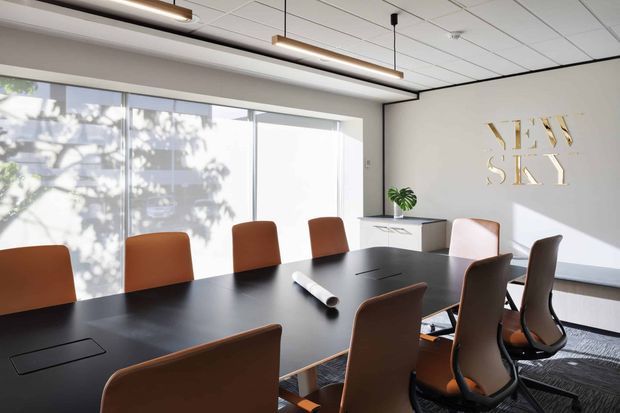Bright contemporary boardroom with a large dark conference table, ergonomic brown and black office chairs, full-height windows, linear pendant lighting, and a white feature wall displaying metallic “NEW SKY” lettering. A small credenza and plant complete the clean corporate interior.