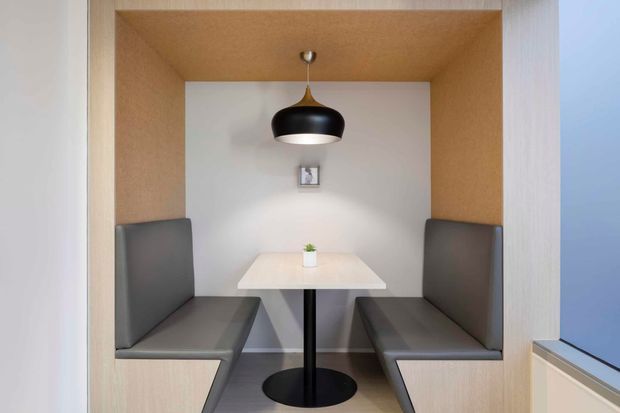 A compact built-in dining nook with two facing upholstered banquette benches, a small white rectangular table, and a central black pendant light. The seating is enclosed within a light timber-framed alcove with warm wood lining, white walls, and grey upholstery.