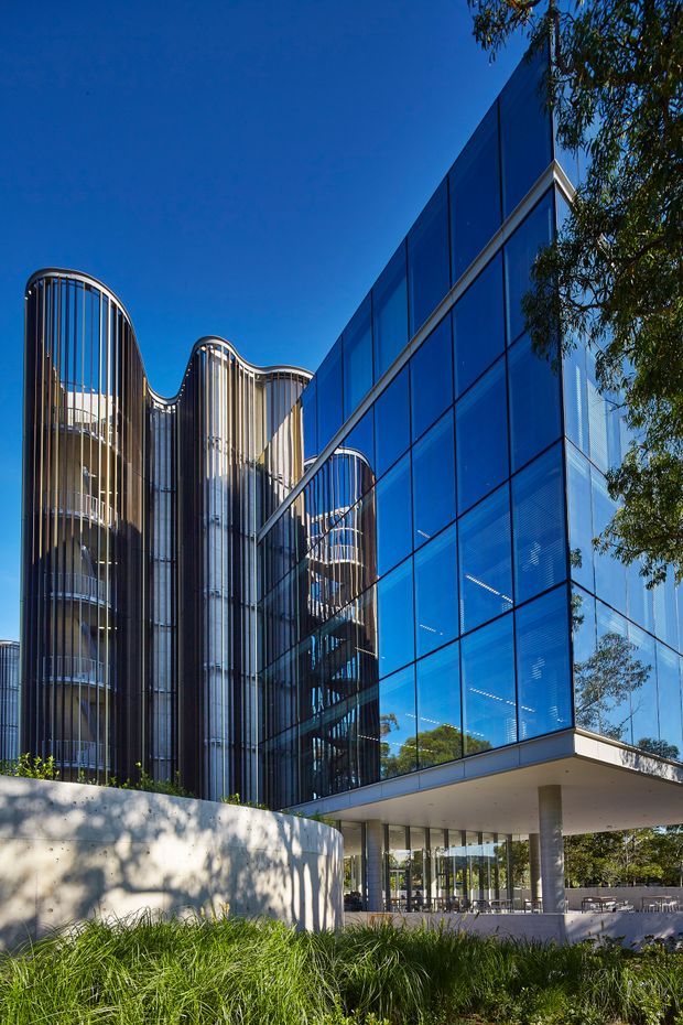 Exterior architectural photograph of a contemporary multi-storey commercial building. The facade combines extensive reflective blue curtain-wall glazing with tall rounded vertical fins and curved glazed forms. A projecting upper volume forms a sheltered entry or terrace supported by slender columns, with landscaped grasses and trees around the base.