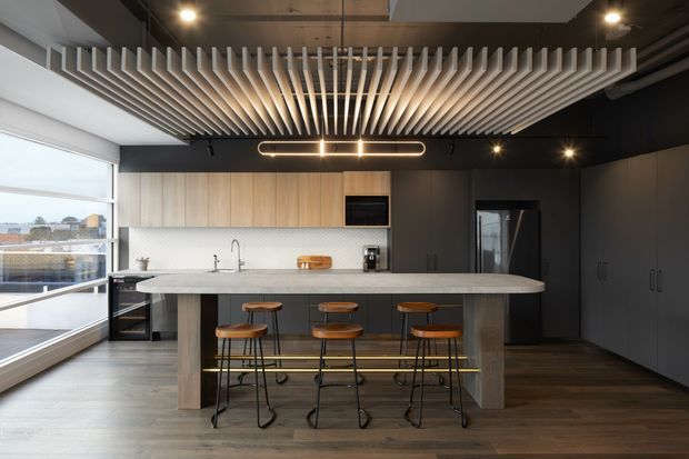 A contemporary kitchen with a large central island and four timber bar stools. The space features dark built-in cabinetry, light timber upper cabinetry, a pale stone island benchtop, wood-look flooring, expansive corner glazing, and a striking ceiling of suspended timber battens with integrated lighting.