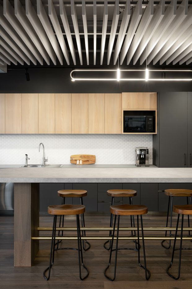A contemporary kitchen with a long island and integrated seating. The space combines dark cabinetry, light timber overhead cabinets, a pale stone-look benchtop, and a white textured splashback. Black appliances and cabinetry contrast with warm timber stools and dark timber flooring. The ceiling features an exposed slatted timber-look structure with integrated linear lighting, creating a refined modern fit-out.