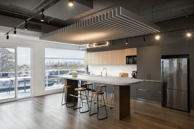 Contemporary open-plan kitchen with a large island, integrated appliances, pale wood and grey cabinetry, and an architectural slatted ceiling feature. Floor-to-ceiling glazing opens to a balcony and provides expansive urban views. Track lighting and dark exposed ceiling services add an industrial influence.