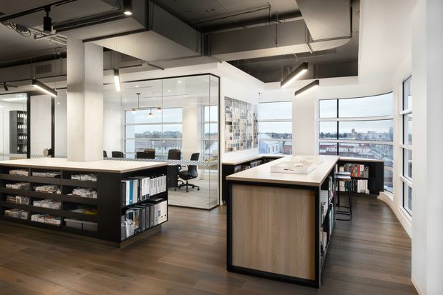 Open-plan contemporary office interior with exposed dark services and ductwork, polished concrete columns, warm timber flooring, extensive glazing, and built-in black shelving and workstations. A central island workstation with integrated storage anchors the space, while a glazed meeting room and city views are visible beyond.