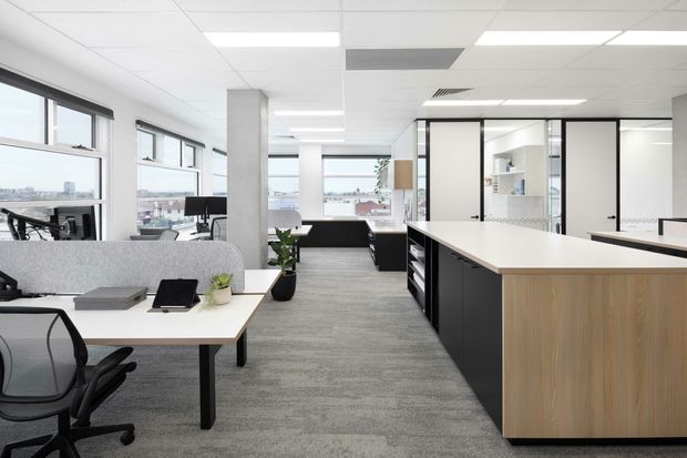 Bright open-plan commercial office interior with shared workstations, large windows, a central kitchen or refreshment island with light wood and black cabinetry, glass-partitioned meeting or utility rooms, grey carpet flooring, white acoustic ceiling tiles, and concrete structural columns. The space has a clean, contemporary corporate fit-out with abundant natural light and views of the surrounding urban area.