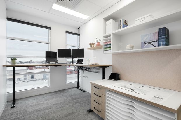 Bright, compact contemporary office with dual workstations positioned beside large windows. The room features white walls, a suspended acoustic ceiling, grey carpet flooring, black-framed glazing, open white shelving, and a built-in drafting or materials desk with drawers and a textured beige backsplash. Multiple monitors, office equipment, and architectural drawings are visible.