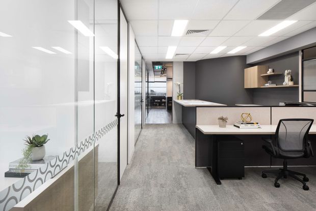 A modern commercial office interior viewed along a central corridor, with glass-partitioned meeting or office rooms on the left and a contemporary workstation on the right. The space features grey carpet flooring, dark charcoal walls, white acoustic ceiling tiles with recessed lighting, black-framed glazing, and light timber open shelving and cabinetry. Minimal desk accessories and a potted plant add restrained decoration.
