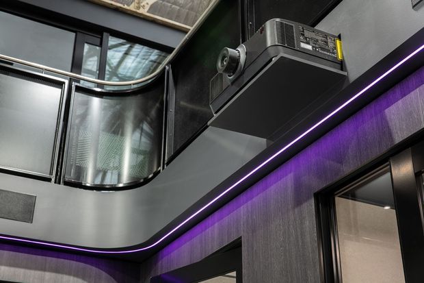 Close-up interior view of a dark, high-tech architectural space with curved black-framed glazing, a projecting upper-level or balcony edge, grey textured wall cladding, and continuous purple LED strip lighting. A ceiling-mounted projector or audiovisual device is visible, suggesting an entertainment or media-focused environment.