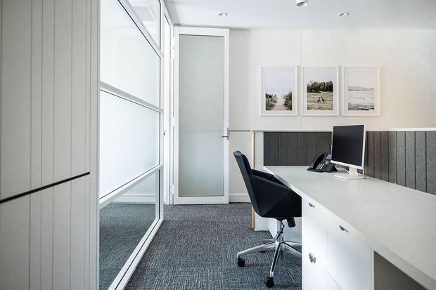 A bright, compact contemporary office workspace with a white built-in desk and storage, charcoal acoustic wall panelling, grey carpet, a black ergonomic office chair, computer monitor and telephone. Full-height internal glazing and a frosted glass door create a light-filled, professional environment, with three framed landscape artworks on the wall.