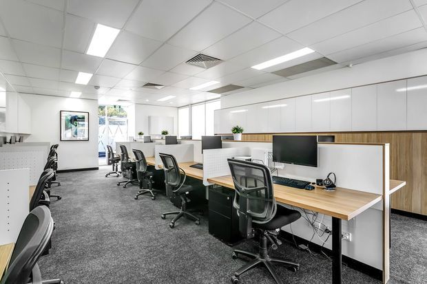 Bright contemporary open-plan office with rows of individual workstations, ergonomic black office chairs, timber-look desks, white partition screens, integrated storage cabinetry, computer monitors, and grey carpet flooring. The space has a suspended white acoustic tile ceiling with recessed fluorescent lighting and large windows providing daylight.