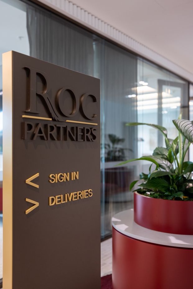 A contemporary commercial reception or entry area featuring a freestanding dark wayfinding sign with raised lettering and gold directional arrows. The sign reads “ROC PARTNERS,” “SIGN IN,” and “DELIVERIES.” Behind it are full-height glazed partitions with sheer curtains, while a large red circular planter with lush green foliage anchors the space.