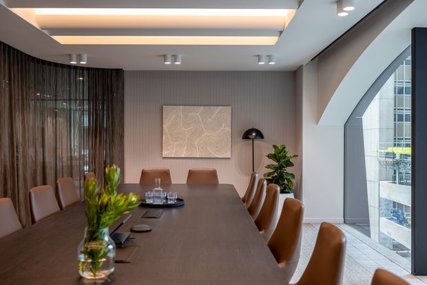 A refined contemporary dining or boardroom space with a long dark wood table surrounded by upholstered tan leather chairs. The room features ribbed grey wall panelling, sheer floor-to-ceiling curtains, recessed ceiling lighting, a sculptural black floor lamp, and a large arched glazed opening overlooking a modern urban building. A floral arrangement and table settings add warmth to the otherwise restrained interior.
