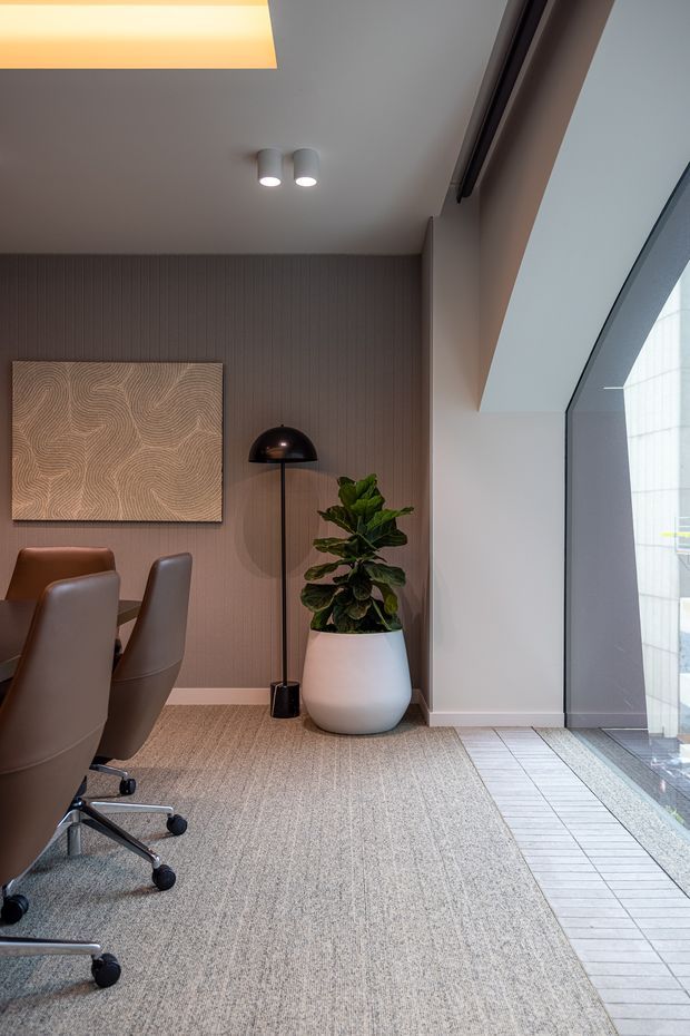 A refined contemporary interior corner, likely a dining or meeting area, with a brown table and upholstered swivel chairs, a dark vertically ribbed feature wall, abstract wood artwork, black dome floor lamp, and a large potted plant. A skylight and full-height glazed opening bring in natural light. The floor combines broadloom carpet with a light tiled strip beside the glazing.