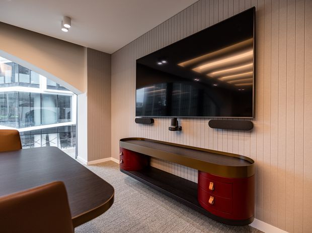 Contemporary media and entertainment area featuring a wall-mounted television, integrated soundbar and speakers, and a curved dark timber console with glossy red drawer units. The room has vertically panelled walls, carpet flooring, recessed and concealed linear lighting, and large glazed openings overlooking an adjacent modern building.