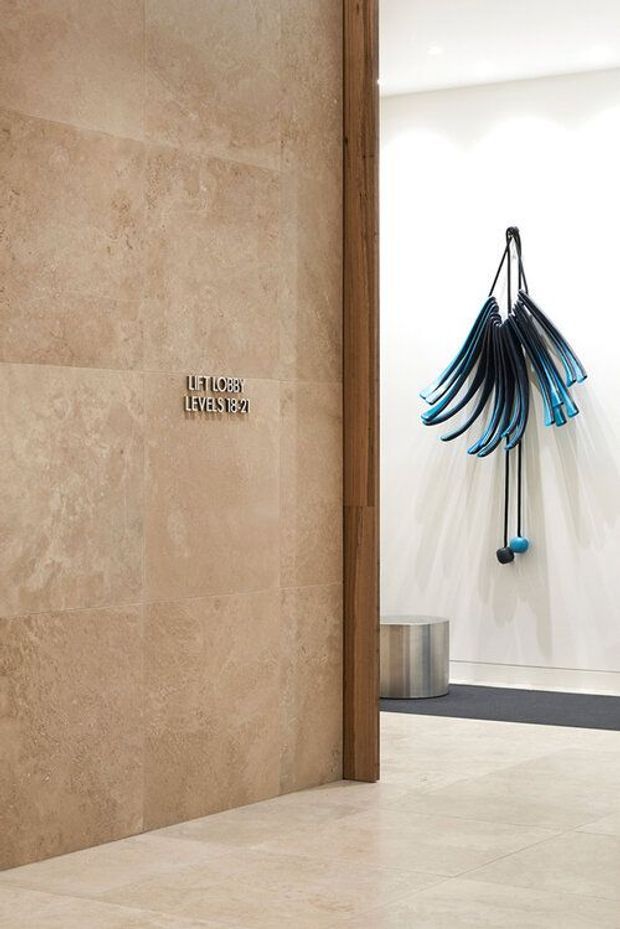 A refined interior lobby or corridor with large beige stone wall and matching tiled floor. A timber-framed opening leads to a bright white foyer featuring a sculptural blue and black wall-mounted artwork, a dark entry mat, and a small metal bin. A small sign identifies lift lobby levels 18–21.