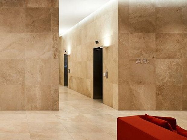 A refined lobby or circulation space clad extensively in large-format warm beige stone tiles, including the walls and floor. Two dark lift doors are set into the stone-clad wall, with discreet linear lighting at the ceiling edge. A vivid red upholstered lounge chair is partially visible in the foreground.