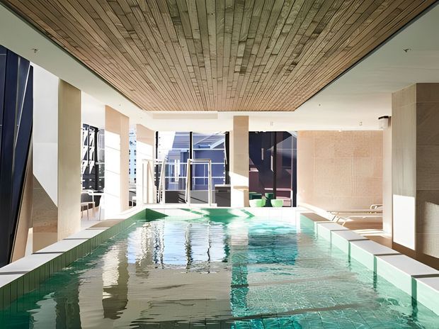 A bright, contemporary indoor swimming pool beneath a large recessed ceiling lined with narrow timber boards. The pool is surrounded by pale tiled surfaces, square structural columns, tall glazed openings, and lounge seating, creating a clean resort-like interior with strong natural light and turquoise water.