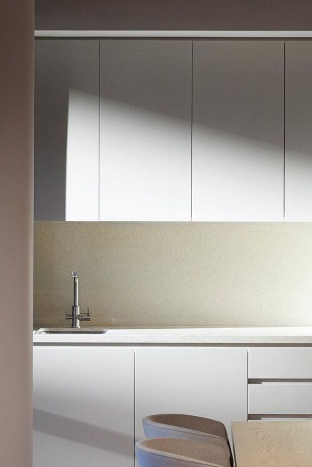 A compact, minimalist kitchen with matte white full-height upper and lower cabinetry, a pale speckled stone benchtop and matching splashback, and a stainless-steel tap and sink. Warm sunlight creates strong diagonal shadows across the cabinetry. Two upholstered stools sit at a small peninsula or dining counter in the foreground.