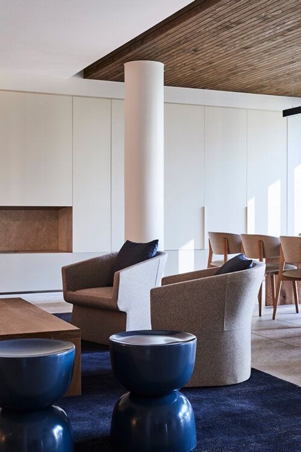 A refined open-plan living and dining area with white built-in cabinetry, a warm timber-lined recessed ceiling, a large cylindrical white column, upholstered armchairs, a wooden dining table and chairs, and glossy deep-blue sculptural side tables on a dark blue rug. The space is bright with a restrained contemporary palette and strong natural light.