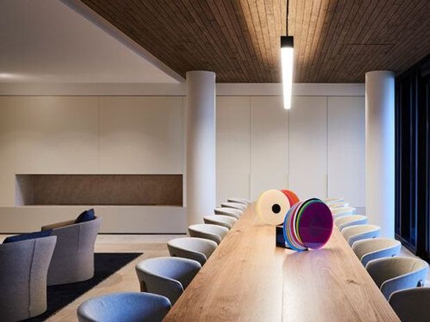 Contemporary dining and meeting space dominated by a long light-wood table surrounded by upholstered chairs. The room features white cylindrical columns, integrated pale wall cabinetry with a recessed darker feature panel, a dark timber slatted ceiling, and full-height dark-framed glazing at the right. Sculptural colourful objects sit on the table.
