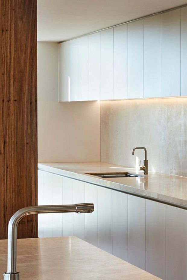 A refined minimalist kitchen with continuous white vertical-panel cabinetry, pale stone benchtops, an integrated sink, and brushed metal tapware. A warm vertical timber-clad wall adds contrast, while concealed under-cabinet lighting illuminates the pale textured splashback. The foreground includes a kitchen island with a sculptural curved faucet.