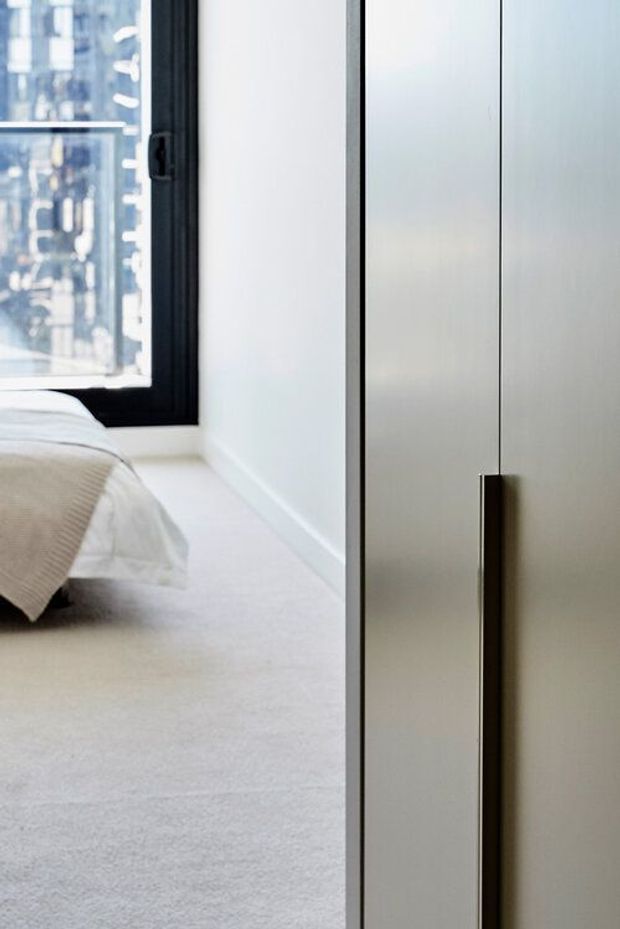 A close-up view of a minimalist bedroom showing the edge of a neatly made bed, pale carpeted flooring, white walls, a large black-framed window, and a tall built-in wardrobe with sleek pale doors and a long dark vertical handle. The image has a clean, understated contemporary aesthetic with soft natural light and an urban view outside.