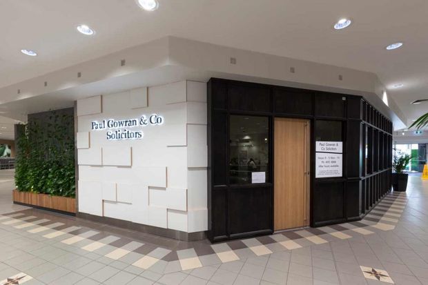 A contemporary commercial solicitor's office entrance in an indoor public lobby. The frontage combines a white geometric feature wall with signage, a dark timber or black-framed office enclosure, a warm timber entry door, extensive glazing, recessed ceiling lights, and a tiled floor with a patterned border. A vertical green wall and lobby circulation areas are visible at the sides.