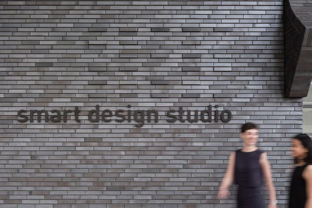 Exterior architectural photograph of a contemporary commercial building entrance or facade, featuring a dark grey horizontally laid brick wall with raised lettering reading “smart design studio.” Two blurred pedestrians appear in the foreground, and a projecting dark architectural volume is visible at the upper right.