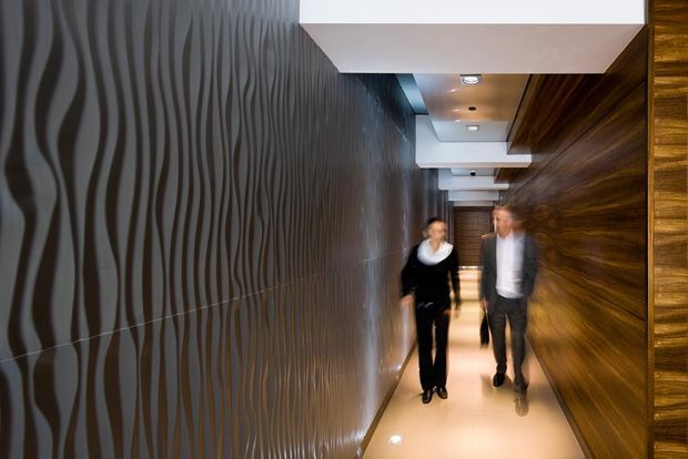 A narrow, modern interior corridor with dramatic dark textured wall panels on the left, warm dark timber wall cladding on the right, and a glossy light-coloured floor. Recessed ceiling lights and repeated white ceiling bulkheads lead toward two blurred people walking through the passage.