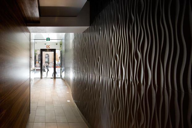 A narrow, dimly lit commercial or building entry corridor leading to a glazed street entrance. The right wall is dominated by a dramatic three-dimensional undulating relief surface in dark tones, while the left wall has dark timber-like wall panels. Large rectangular floor tiles and recessed ceiling panels create a refined contemporary passageway.