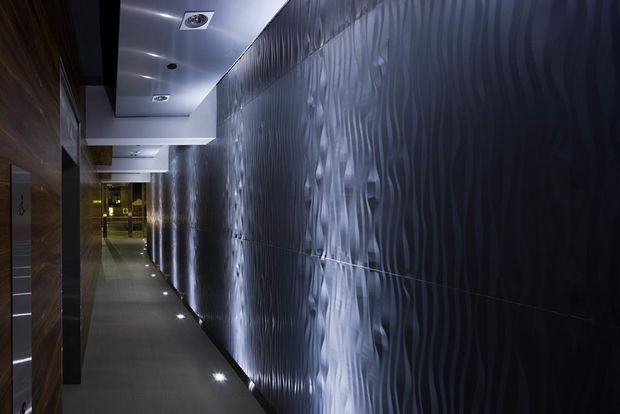 A narrow, dimly lit contemporary corridor with a dramatic dark textured feature wall illuminated by repeated low-level uplights. The opposite side has dark timber-toned wall panels and doorways, while recessed ceiling spotlights and a linear arrangement of floor lights lead toward a brighter area at the end.