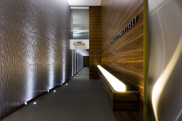 A narrow, dramatic architectural corridor or entry passage with dark grey tiled flooring, a highly textured metallic feature wall on the left, and richly grained dark timber wall panelling on the right. A long integrated timber bench with concealed linear lighting runs beneath raised lettering reading “WILLIAM STREET.” Recessed floor-level lights and reflective surfaces create a sophisticated, gallery-like atmosphere.