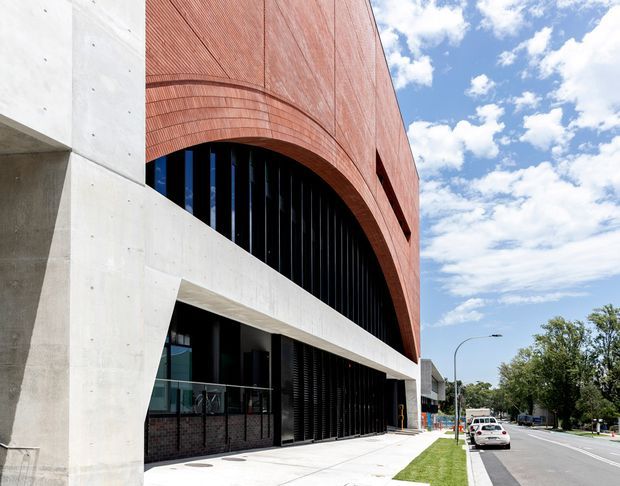 Exterior streetscape view of a contemporary multi-storey building combining a curved red brick facade with broad arched openings, exposed pale concrete structural elements, dark vertical screening, and glazed lower-level frontage. The building sits along an urban/suburban roadway under a bright sky.